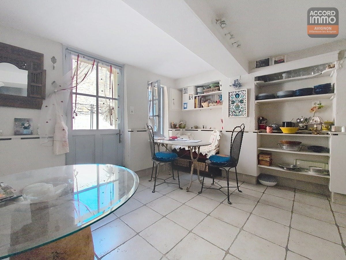 Espace cuisine avec porte d'entrée vitrée peinte en bleu-gris ouvrant sur l'extérieur. Table ronde en marbre, chaises bistrot en fer forgé à assises bleues, étagères murales blanches avec vaisselle et sol carrelé clair.