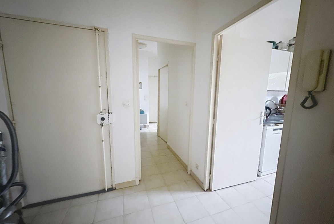 Hall d'entrée avec porte blindée beige équipée de verrous, interphone mural et carrelage clair au sol. Couloir desservant les pièces avec portes blanches ouvertes sur cuisine visible à droite et pièce à vivre au fond, murs blancs et aspirateur rangé près de l'entrée.