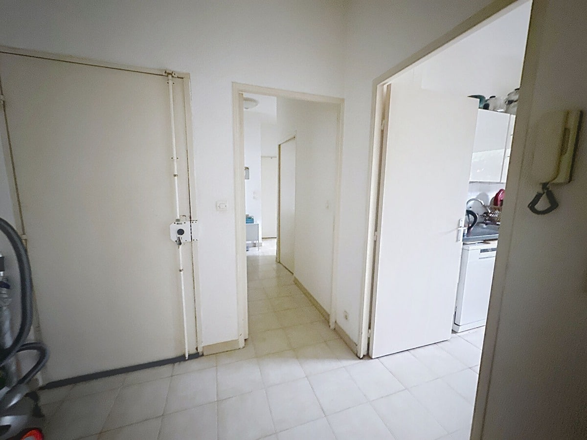 Hall d'entrée avec porte blindée beige équipée de verrous, interphone mural et carrelage clair au sol. Couloir desservant les pièces avec portes blanches ouvertes sur cuisine visible à droite et pièce à vivre au fond, murs blancs et aspirateur rangé près de l'entrée.