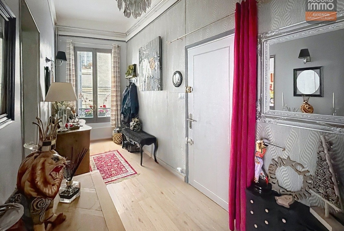 Hall d'entrée avec parquet clair, murs gris à motifs zébrés, grand miroir baroque argenté et rideau fuchsia. Statue de tigre décorative, chaise noire style baroque, petit tapis persan rouge. Fenêtre avec vue sur rue, lustre à pampilles et console avec objets décoratifs.