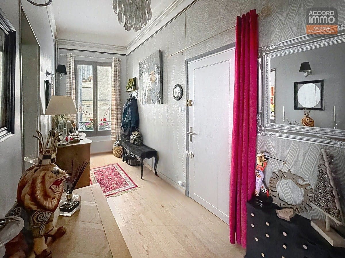 Hall d'entrée avec parquet clair, murs gris à motifs zébrés, grand miroir baroque argenté et rideau fuchsia. Statue de tigre décorative, chaise noire style baroque, petit tapis persan rouge. Fenêtre avec vue sur rue, lustre à pampilles et console avec objets décoratifs.