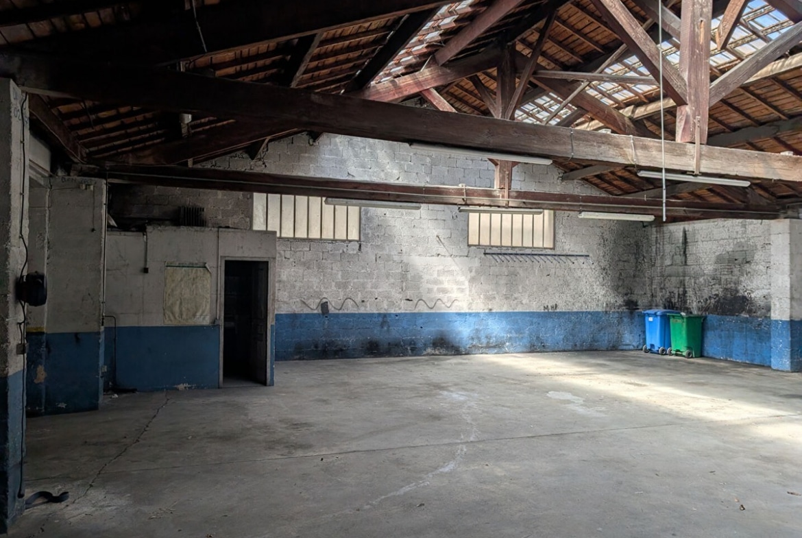 Grand volume avec charpente en bois apparente et toiture traditionnelle laissant filtrer la lumière par des ouvertures. Murs en parpaings gris avec bande bleue peinte en partie basse, sol en béton lissé, poubelles bleue et verte visibles contre le mur du fond, porte métallique sur la gauche.