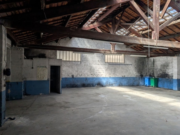 Grand volume avec charpente en bois apparente et toiture traditionnelle laissant filtrer la lumière par des ouvertures. Murs en parpaings gris avec bande bleue peinte en partie basse, sol en béton lissé, poubelles bleue et verte visibles contre le mur du fond, porte métallique sur la gauche.