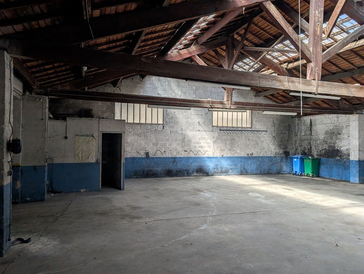 Grand volume avec charpente en bois apparente et toiture traditionnelle laissant filtrer la lumière par des ouvertures. Murs en parpaings gris avec bande bleue peinte en partie basse, sol en béton lissé, poubelles bleue et verte visibles contre le mur du fond, porte métallique sur la gauche.