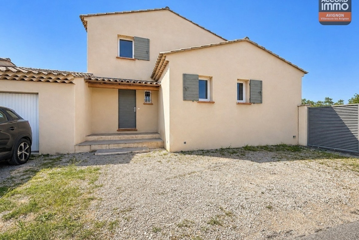 Maison à étage avec enduit beige et volets gris-vert. Entrée avec porte grise et petit auvent en bois, accessible par quelques marches en pierre. Cour en gravier devant la façade.