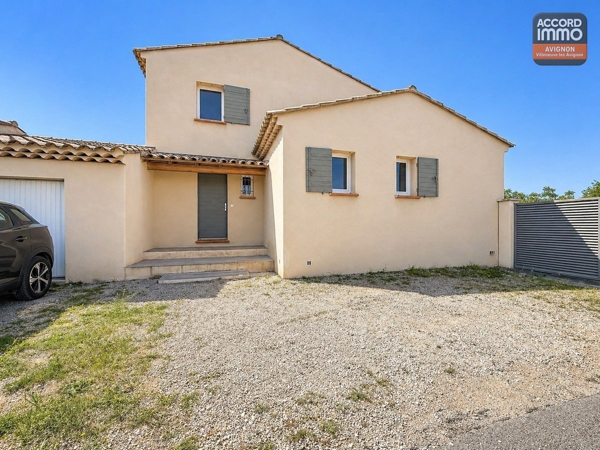 Maison à étage avec enduit beige et volets gris-vert. Entrée avec porte grise et petit auvent en bois, accessible par quelques marches en pierre. Cour en gravier devant la façade.