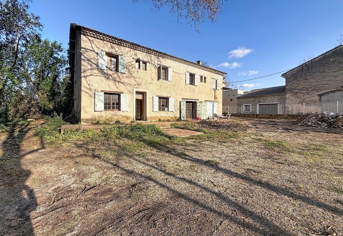 Bâtiment ancien à deux étages avec façade en pierre enduite de couleur ocre et toiture en tuiles canal. Multiples fenêtres à petits carreaux encadrées de volets en bois vert pâle et blanc, portes d'entrée en bois, cour en terre battue avec végétation éparse au premier plan.