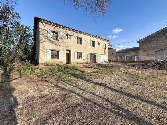 Bâtiment ancien à deux étages avec façade en pierre enduite de couleur ocre et toiture en tuiles canal. Multiples fenêtres à petits carreaux encadrées de volets en bois vert pâle et blanc, portes d'entrée en bois, cour en terre battue avec végétation éparse au premier plan.