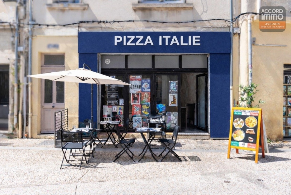 Devanture de pizzeria avec enseigne "Pizza Italie" sur fond bleu marine et lettrage blanc. Terrasse extérieure composée de trois tables métalliques noires avec chaises pliantes sous un parasol écru, chevalet publicitaire présentant des spécialités italiennes sur le trottoir pavé.