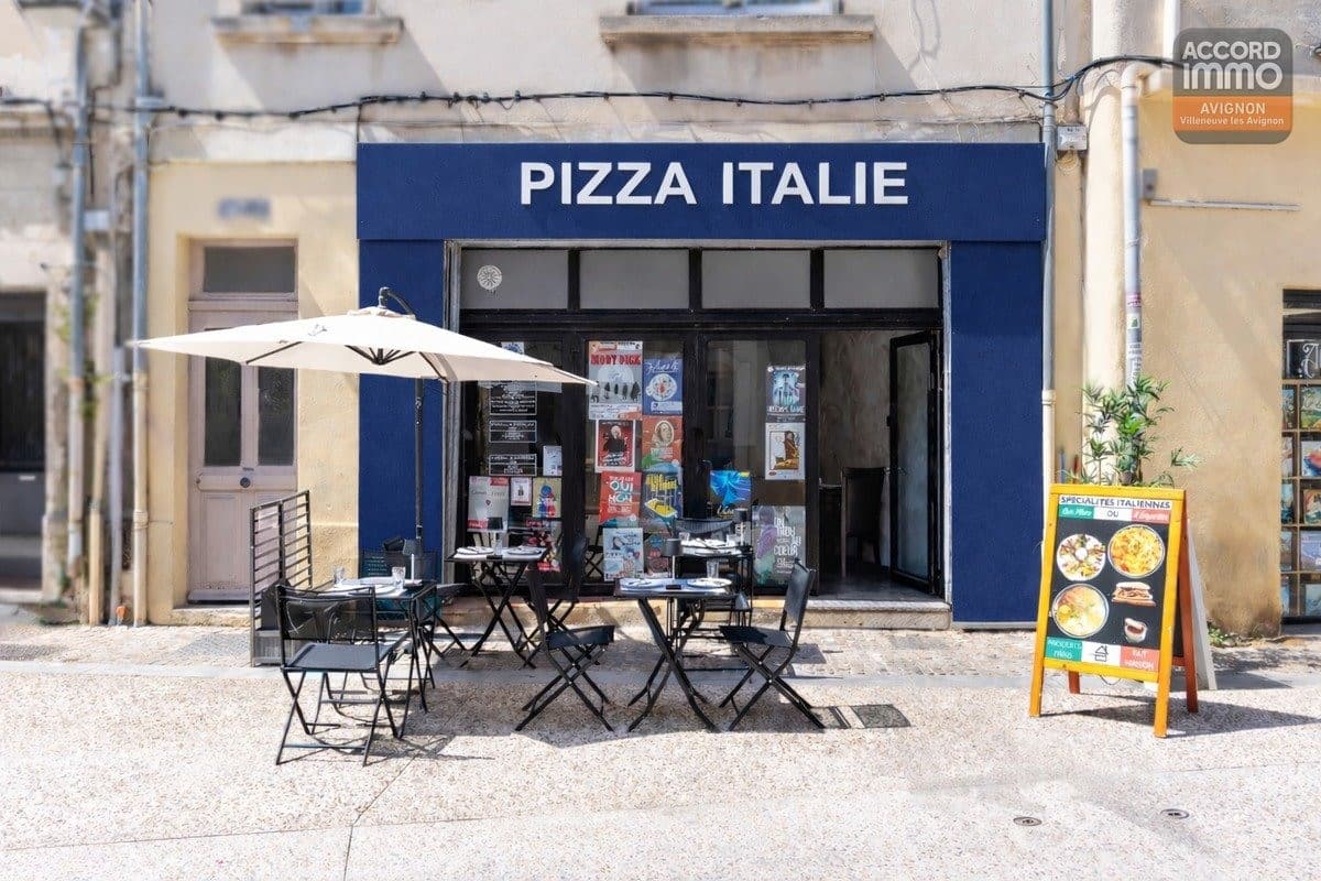 Devanture de pizzeria avec enseigne "Pizza Italie" sur fond bleu marine et lettrage blanc. Terrasse extérieure composée de trois tables métalliques noires avec chaises pliantes sous un parasol écru, chevalet publicitaire présentant des spécialités italiennes sur le trottoir pavé.