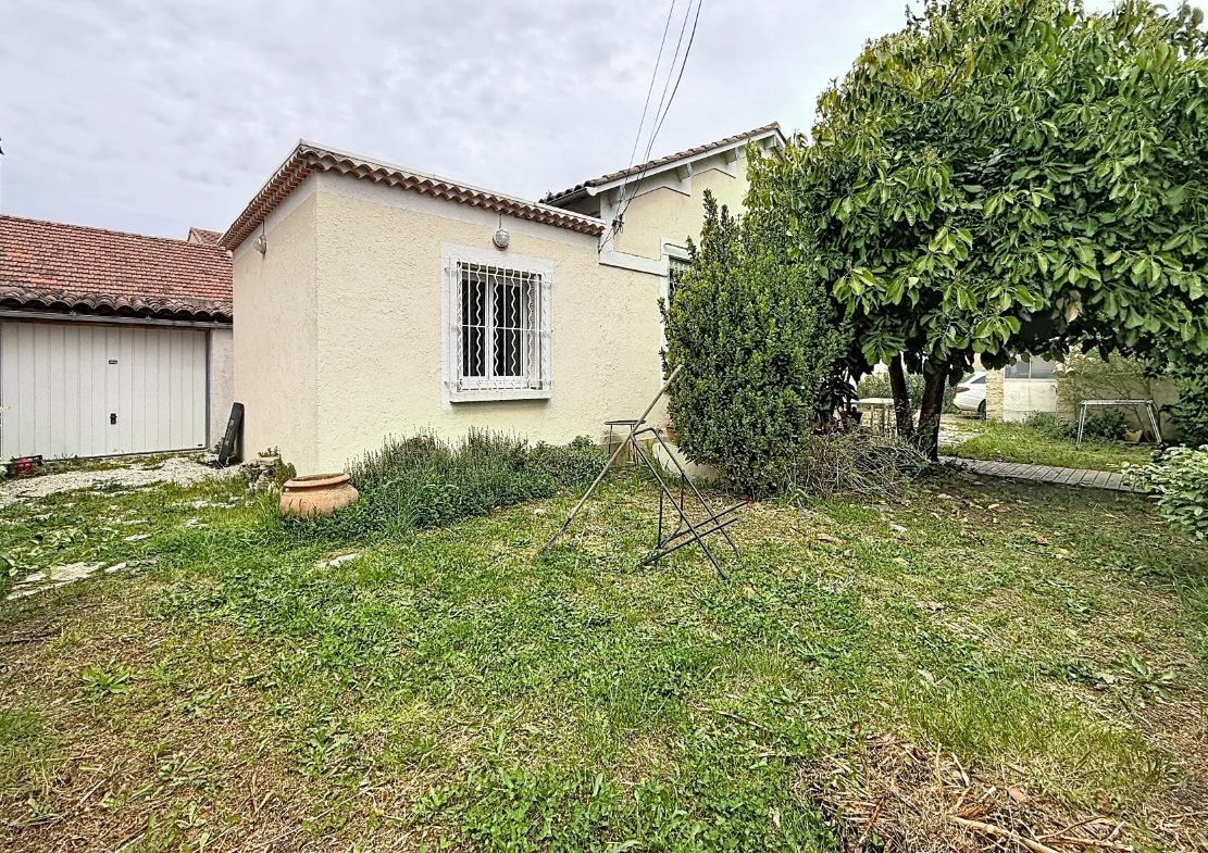 Vue sur le jardin enherbé d'une maison avec façade enduite beige, fenêtre protégée par des barreaux blancs et garage attenant avec porte basculante blanche et toiture en tuiles canal. Terrain planté de haies, arbustes taillés, un grand arbre et un étendoir métallique au centre de la pelouse.