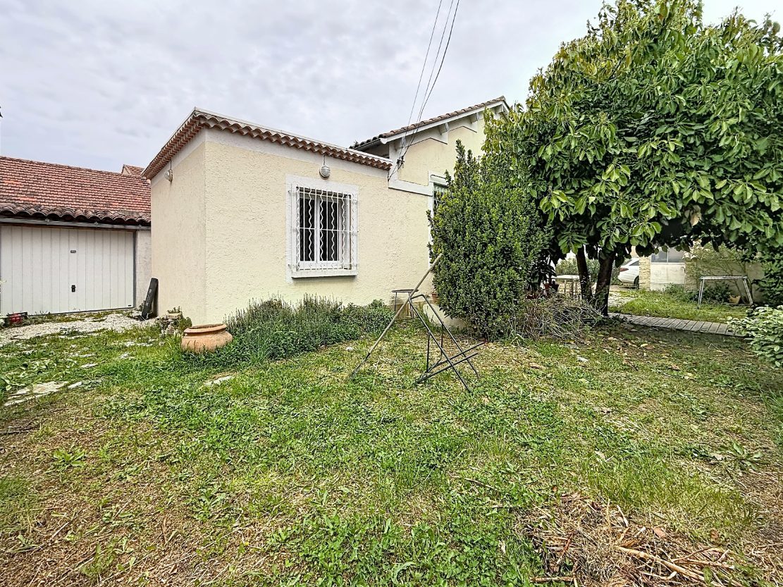 Vue sur le jardin enherbé d'une maison avec façade enduite beige, fenêtre protégée par des barreaux blancs et garage attenant avec porte basculante blanche et toiture en tuiles canal. Terrain planté de haies, arbustes taillés, un grand arbre et un étendoir métallique au centre de la pelouse.