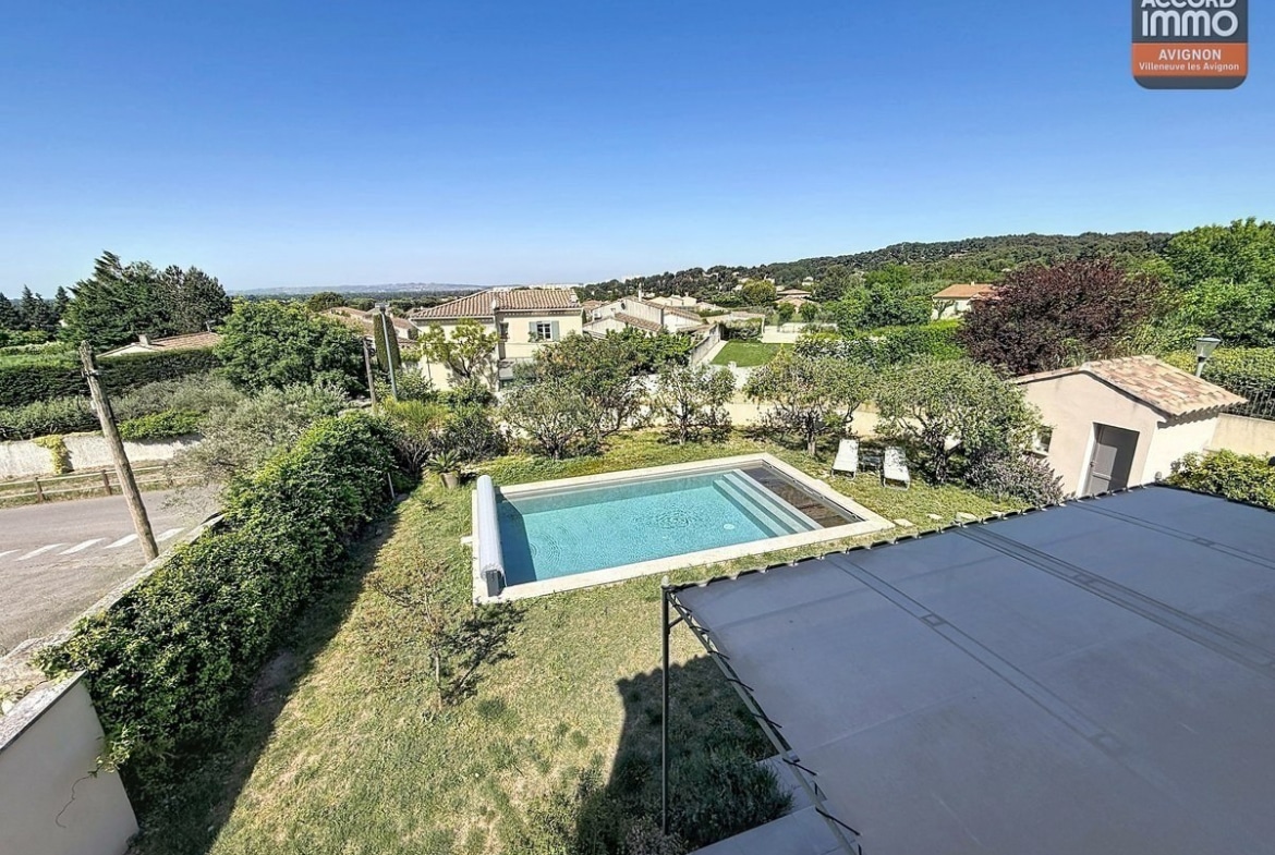 Vue plongeante sur le jardin avec piscine rectangulaire et pelouse. Terrasse couverte visible à droite, cabanon au fond du terrain. Environnement résidentiel verdoyant avec collines en arrière-plan.