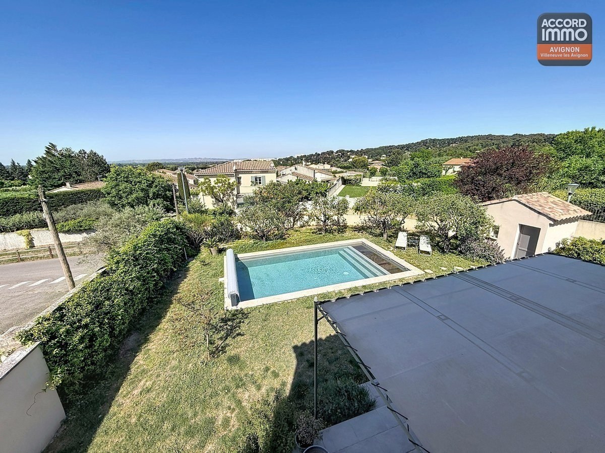 Vue plongeante sur le jardin avec piscine rectangulaire et pelouse. Terrasse couverte visible à droite, cabanon au fond du terrain. Environnement résidentiel verdoyant avec collines en arrière-plan.