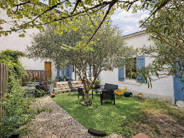 Jardin clos avec olivier mature au centre, pelouse verte et salon de jardin avec fauteuils noirs et chaise jaune. Façade blanche avec volets bleus provençaux, escalier en pierre menant à terrasse et haie végétale en bordure.