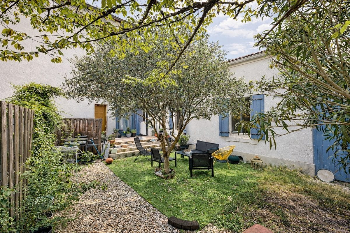 Jardin clos avec olivier mature au centre, pelouse verte et salon de jardin avec fauteuils noirs et chaise jaune. Façade blanche avec volets bleus provençaux, escalier en pierre menant à terrasse et haie végétale en bordure.