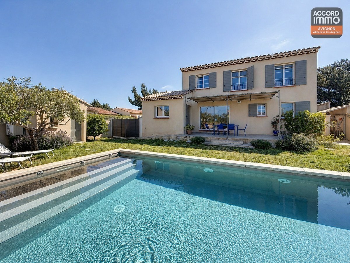 Piscine rectangulaire avec margelles en pierre et eau turquoise au centre d'un jardin engazonné. Terrasse couverte avec pergola en toile et mobilier de jardin bleu devant la maison à étage.