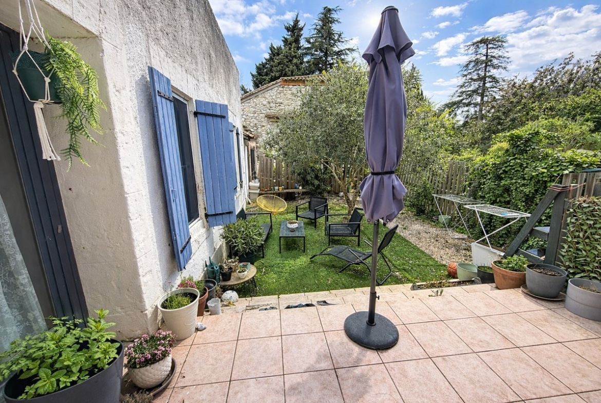Jardin privatif avec pelouse verte, olivier central et salon de jardin composé de fauteuils noirs. Terrasse carrelée en terre cuite avec parasol gris fermé, pots de plantes et volets bleus provençaux sur façade blanche.