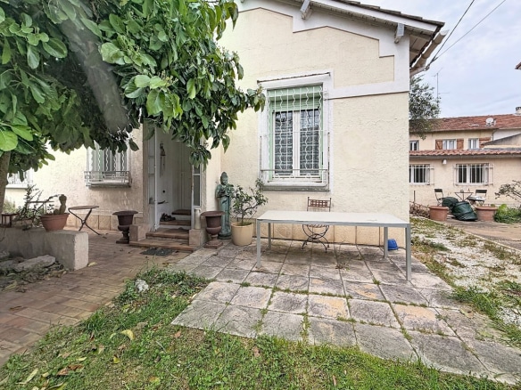 Façade d'une maison aux murs enduits beige avec fenêtres équipées de grilles en fer forgé blanc et porte d'entrée accessible par quelques marches. Jardin composé d'une terrasse dallée en pierre, d'une table rectangulaire blanche, de pots en terre cuite, d'une statue décorative et d'un grand arbre feuillu sur la gauche.