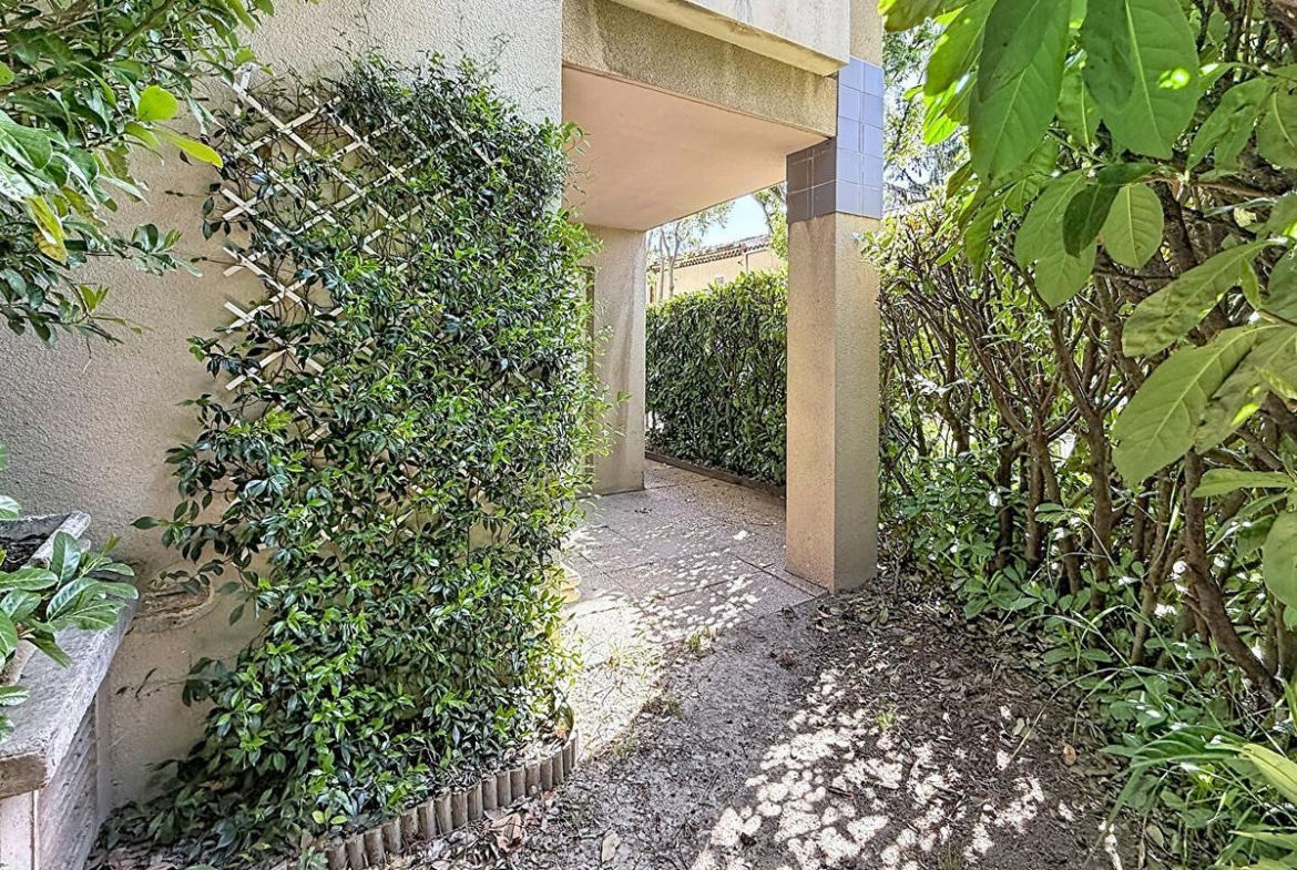 Allée extérieure bordée de végétation dense avec jasmin grimpant sur treillage blanc contre mur crépi beige. Passage couvert menant vers un jardin privatif avec haies taillées et arbustes, sol en terre battue parsemé de feuilles mortes.