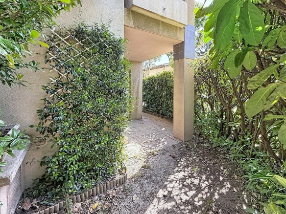 Allée extérieure bordée de végétation dense avec jasmin grimpant sur treillage blanc contre mur crépi beige. Passage couvert menant vers un jardin privatif avec haies taillées et arbustes, sol en terre battue parsemé de feuilles mortes.