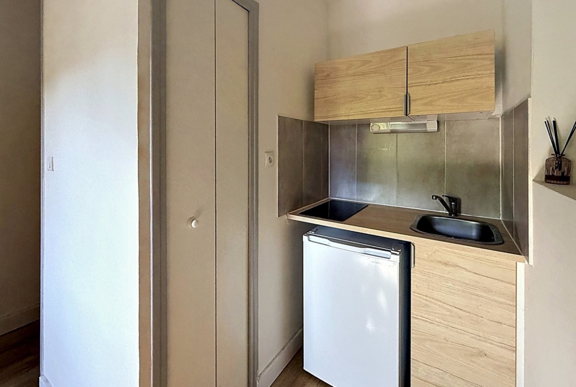 Kitchenette compacte avec meuble haut en bois clair, plaques de cuisson intégrées et évier noir. Réfrigérateur blanc encastré sous le plan de travail, crédence métallique grise, porte de placard à gauche.