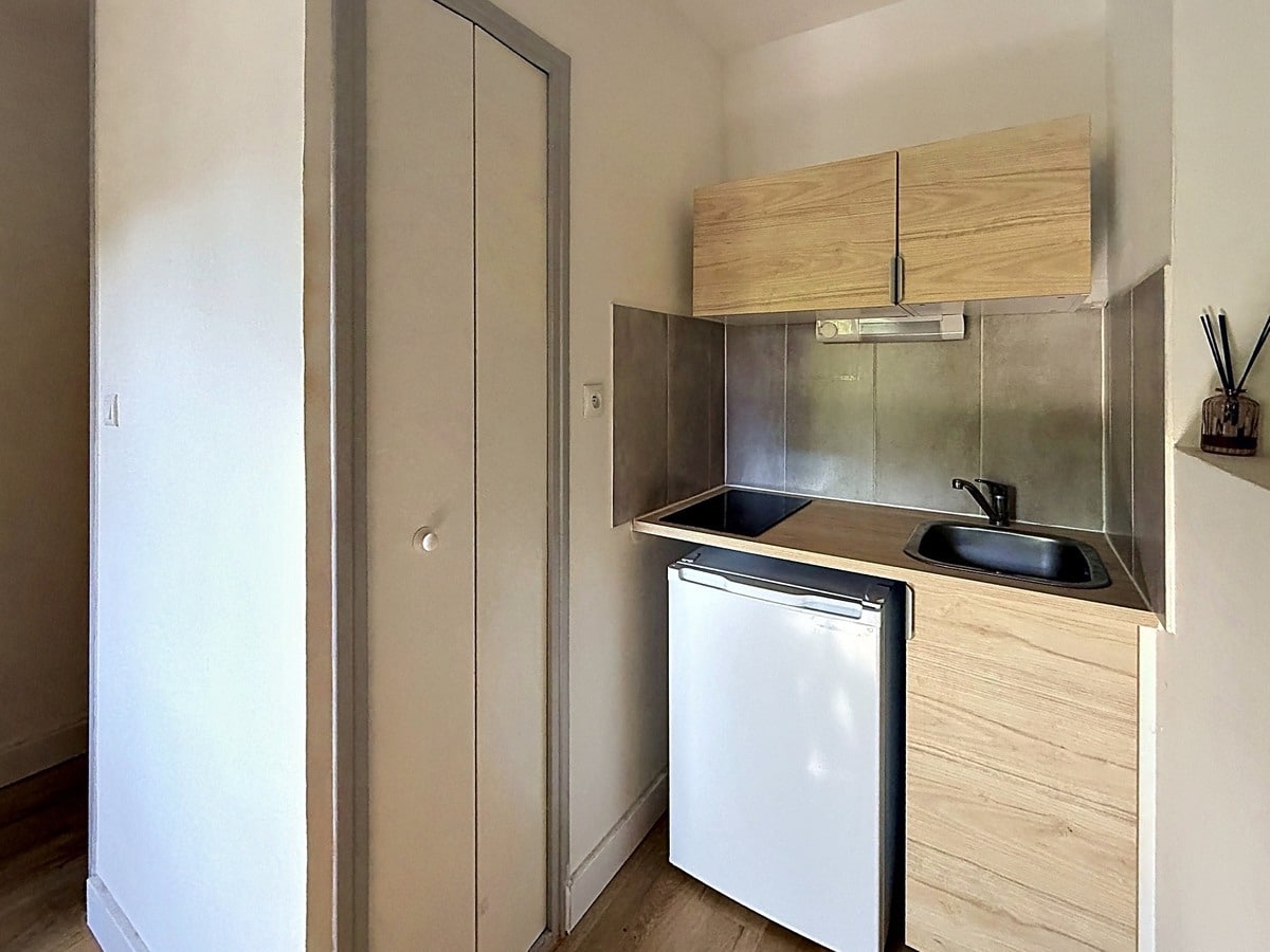 Kitchenette compacte avec meuble haut en bois clair, plaques de cuisson intégrées et évier noir. Réfrigérateur blanc encastré sous le plan de travail, crédence métallique grise, porte de placard à gauche.