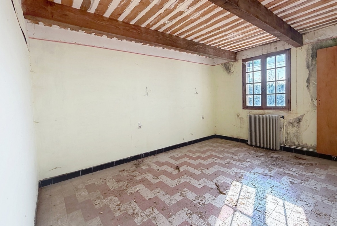 Pièce vide au sol carrelé rose et blanc en motif chevron, avec plinthes bleu foncé et murs blanc cassé présentant des traces d'humidité. Plafond traditionnel avec poutres en bois apparentes et lattis, fenêtre à petits carreaux avec cadre en bois brun et radiateur en fonte sous la fenêtre.