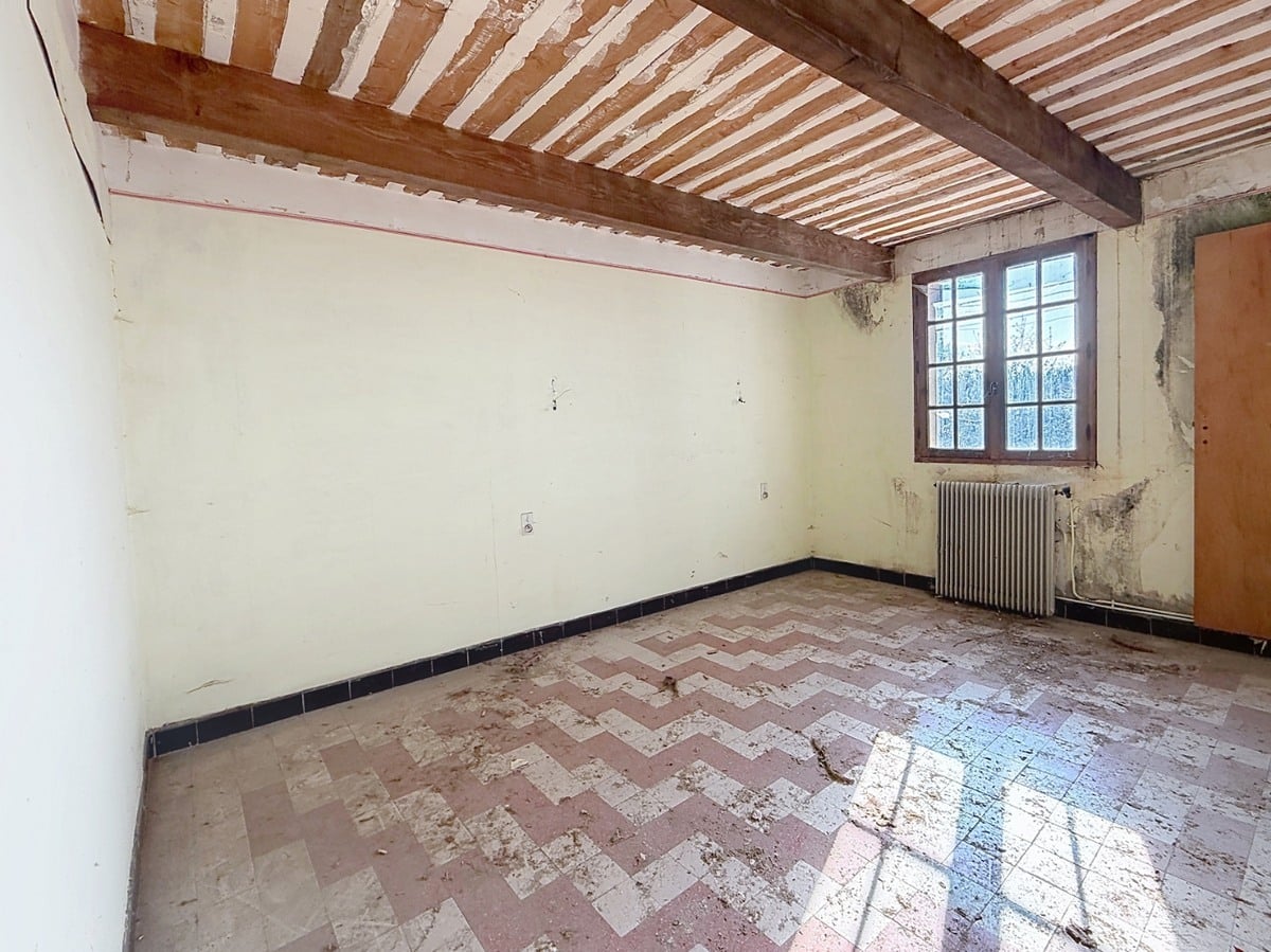 Pièce vide au sol carrelé rose et blanc en motif chevron, avec plinthes bleu foncé et murs blanc cassé présentant des traces d'humidité. Plafond traditionnel avec poutres en bois apparentes et lattis, fenêtre à petits carreaux avec cadre en bois brun et radiateur en fonte sous la fenêtre.