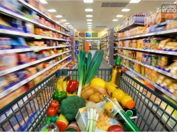 Vue depuis un caddie de supermarché rempli de légumes frais, broccoli, poireaux, poivrons rouges, pommes de terre, bouteilles d'huile et jus d'orange. Les allées du magasin présentent des rayonnages garnis de produits alimentaires variés de part et d'autre, avec un éclairage fluorescent au plafond et un sol carrelé beige.