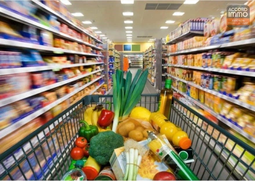 Vue depuis un caddie de supermarché rempli de légumes frais, broccoli, poireaux, poivrons rouges, pommes de terre, bouteilles d'huile et jus d'orange. Les allées du magasin présentent des rayonnages garnis de produits alimentaires variés de part et d'autre, avec un éclairage fluorescent au plafond et un sol carrelé beige.