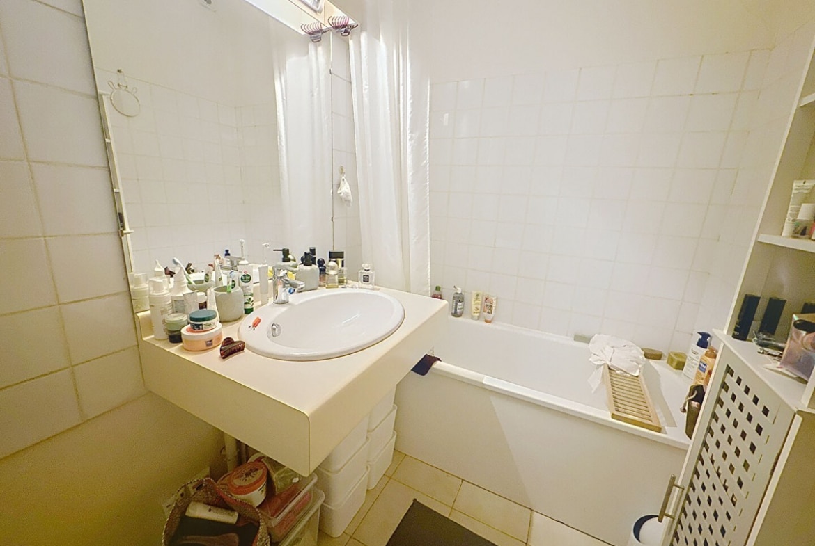 Salle de bains carrelée en blanc avec baignoire équipée d'un rideau de douche transparent, vasque encastrée avec grand miroir et éclairage néon. Nombreux produits de toilette sur le plan vasque, meuble de rangement blanc ajouré et tapis de bain noir au sol.