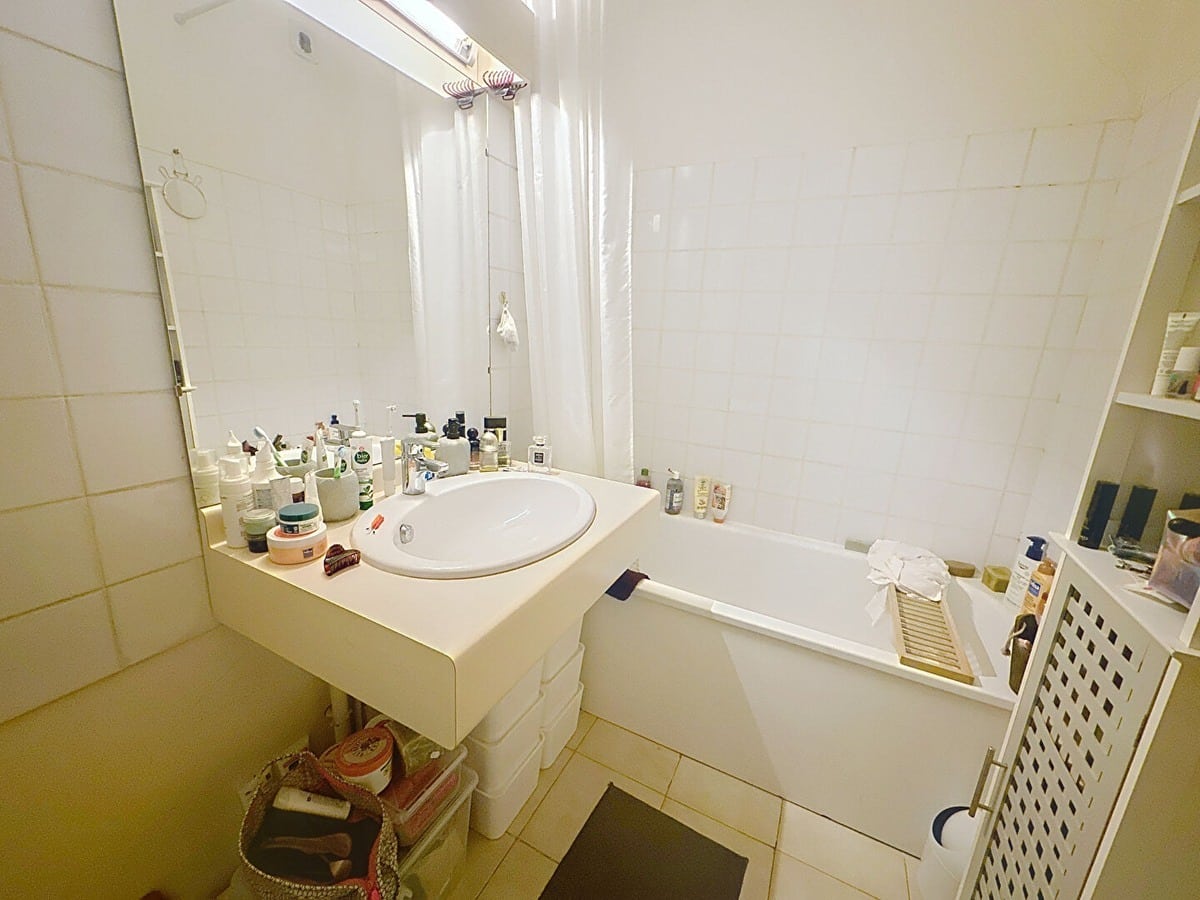 Salle de bains carrelée en blanc avec baignoire équipée d'un rideau de douche transparent, vasque encastrée avec grand miroir et éclairage néon. Nombreux produits de toilette sur le plan vasque, meuble de rangement blanc ajouré et tapis de bain noir au sol.