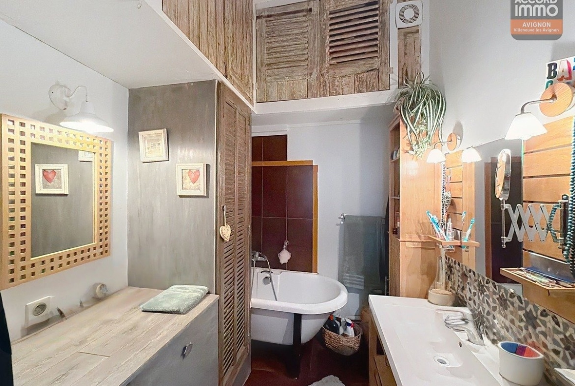 Salle de bains avec baignoire sur pieds blancs dans alcôve carrelée bordeaux, placards en bois clair à persiennes jusqu'au plafond. Vasque blanche sur meuble bois, miroir doré tressé, mur en béton ciré gris. Sol rouge, rangements en bois et plantes vertes décoratives.
