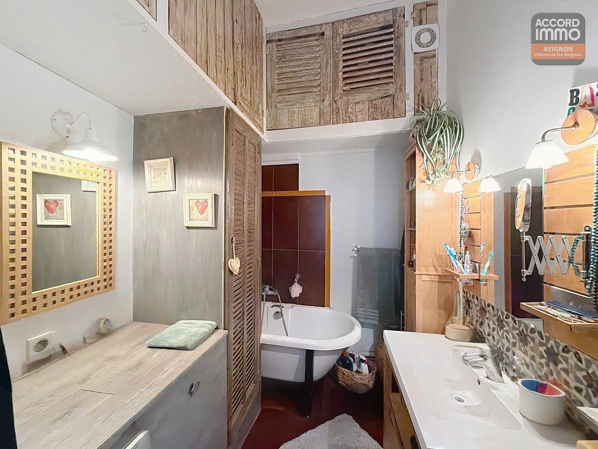 Salle de bains avec baignoire sur pieds blancs dans alcôve carrelée bordeaux, placards en bois clair à persiennes jusqu'au plafond. Vasque blanche sur meuble bois, miroir doré tressé, mur en béton ciré gris. Sol rouge, rangements en bois et plantes vertes décoratives.