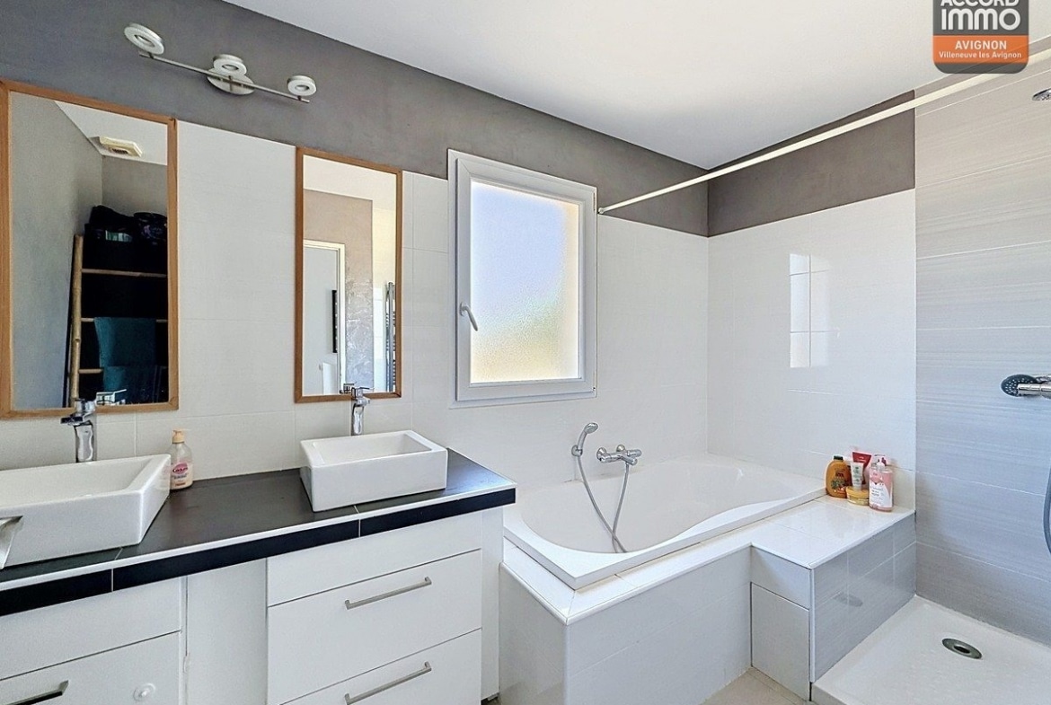 Salle de bains avec double vasque blanche sur meuble, plan de travail noir et miroirs encadrés bois. Baignoire d'angle blanche et cabine de douche à droite, carrelage mural blanc et gris.