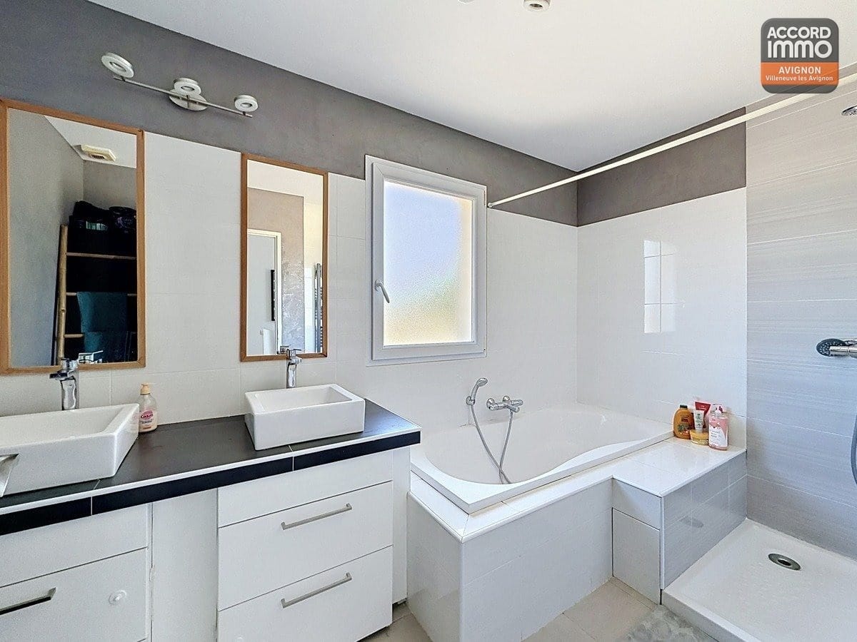 Salle de bains avec double vasque blanche sur meuble, plan de travail noir et miroirs encadrés bois. Baignoire d'angle blanche et cabine de douche à droite, carrelage mural blanc et gris.