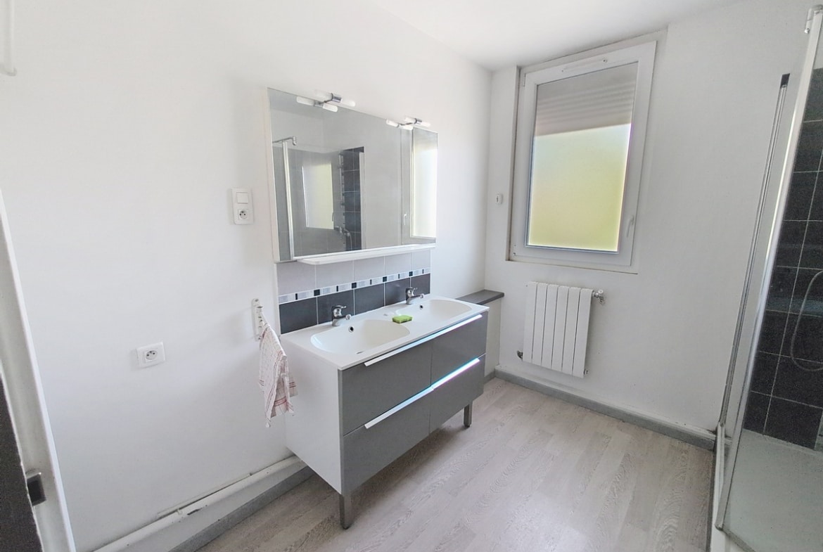 Salle de bains avec meuble double vasque gris et blanc, miroir éclairé et carrelage mural gris anthracite. Cabine de douche vitrée à droite, fenêtre avec volet roulant et radiateur blanc.