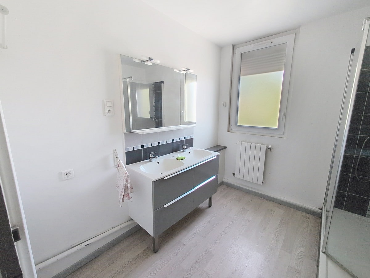 Salle de bains avec meuble double vasque gris et blanc, miroir éclairé et carrelage mural gris anthracite. Cabine de douche vitrée à droite, fenêtre avec volet roulant et radiateur blanc.