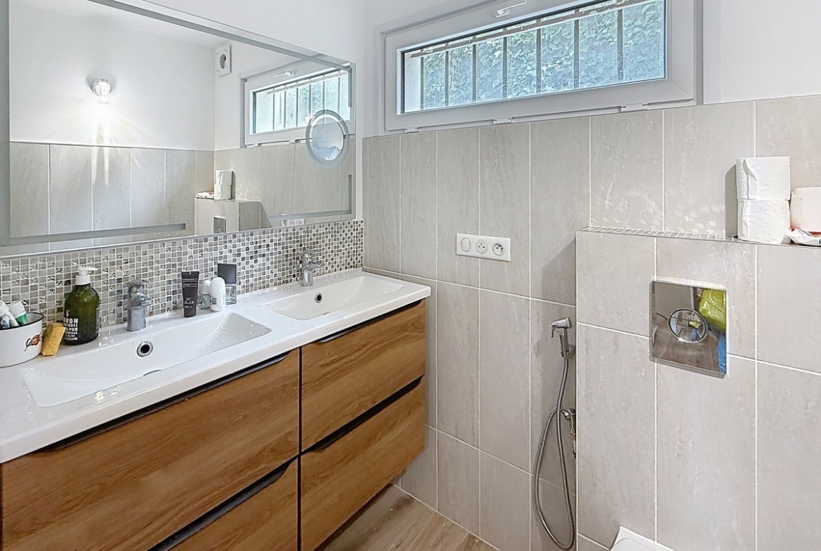 Salle de bains carrelée en tons gris avec meuble vasque en bois et double lavabo blanc. Douche italienne avec robinetterie encastrée et fenêtre à barreaux en partie haute.