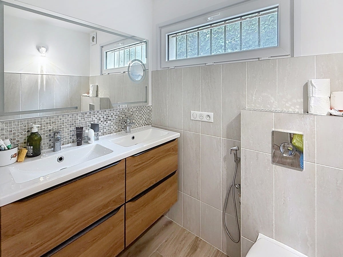 Salle de bains carrelée en tons gris avec meuble vasque en bois et double lavabo blanc. Douche italienne avec robinetterie encastrée et fenêtre à barreaux en partie haute.