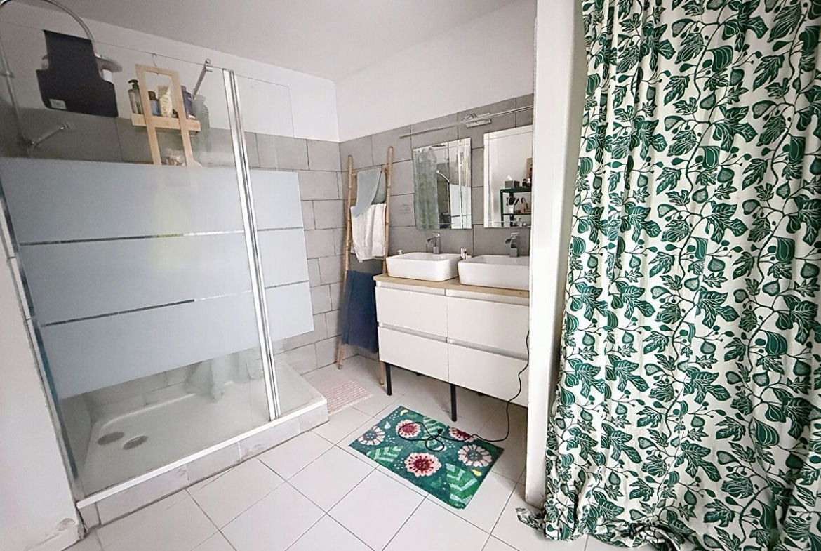 Salle de bains avec cabine de douche vitrée, carrelage mural gris rectangulaire et sol carrelé blanc. Meuble vasque blanc suspendu avec double vasque ronde, miroir et échelle décorative en bambou pour serviettes.