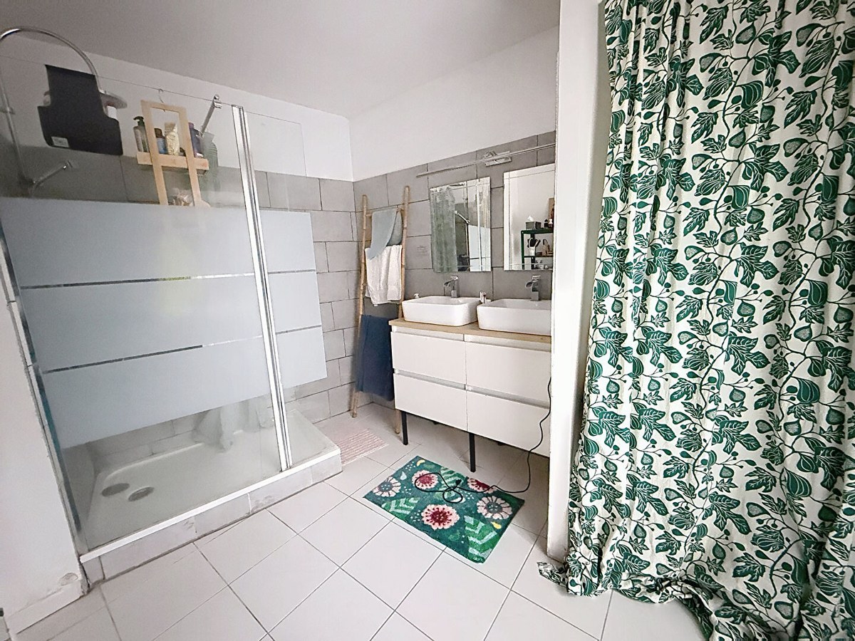 Salle de bains avec cabine de douche vitrée, carrelage mural gris rectangulaire et sol carrelé blanc. Meuble vasque blanc suspendu avec double vasque ronde, miroir et échelle décorative en bambou pour serviettes.