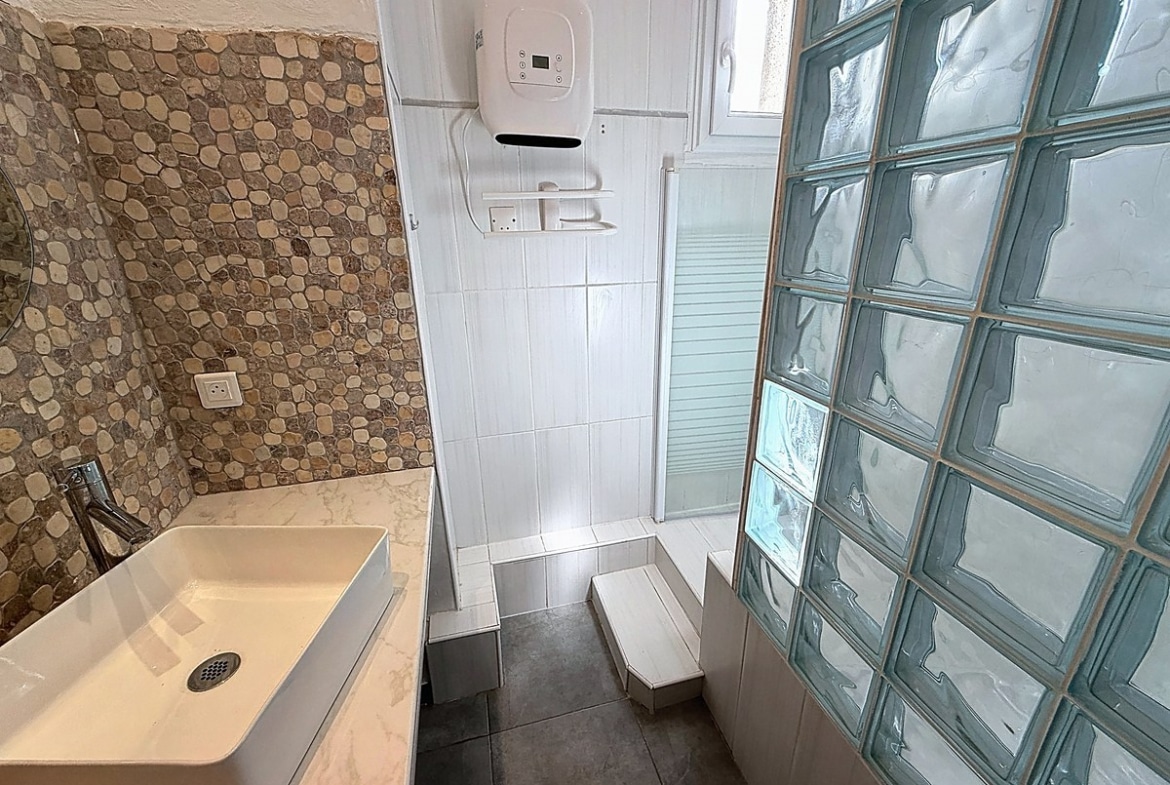 Salle de bains avec vasque rectangulaire blanche sur plan marbré et mur en galets naturels. Douche à l'italienne avec paroi vitrée, cloison en pavés de verre translucides et chauffe-eau mural blanc.