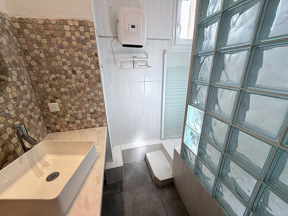Salle de bains avec vasque rectangulaire blanche sur plan marbré et mur en galets naturels. Douche à l'italienne avec paroi vitrée, cloison en pavés de verre translucides et chauffe-eau mural blanc.