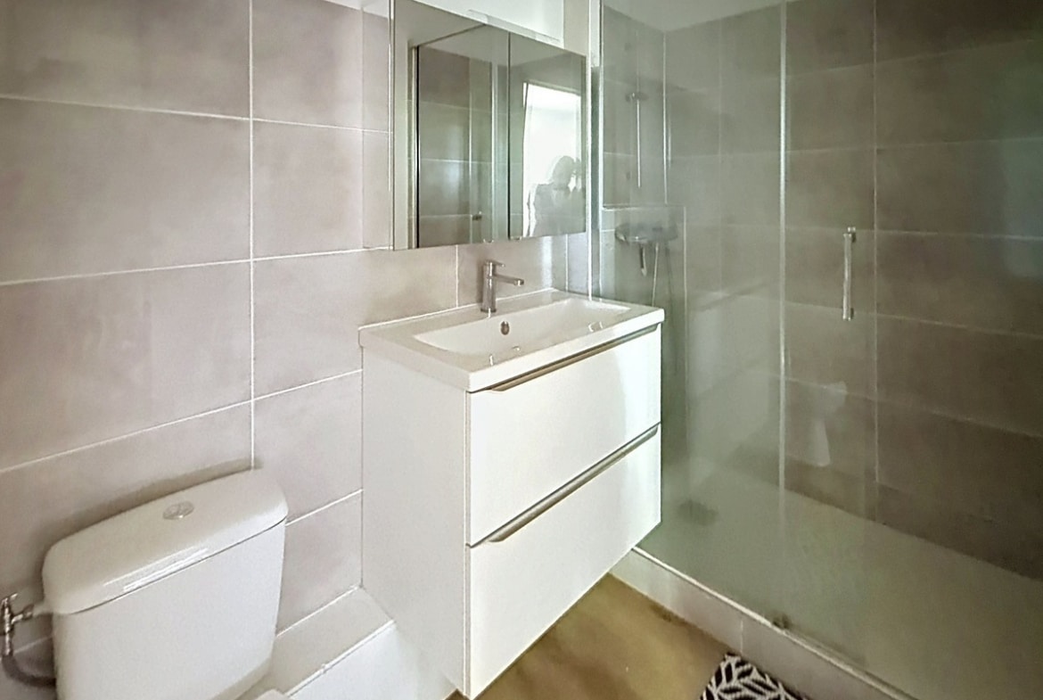 Salle d'eau avec carrelage gris clair, meuble vasque blanc suspendu à deux tiroirs et miroir rectangulaire. Cabine de douche avec paroi vitrée et porte coulissante, WC blanc à gauche, sol en parquet clair.