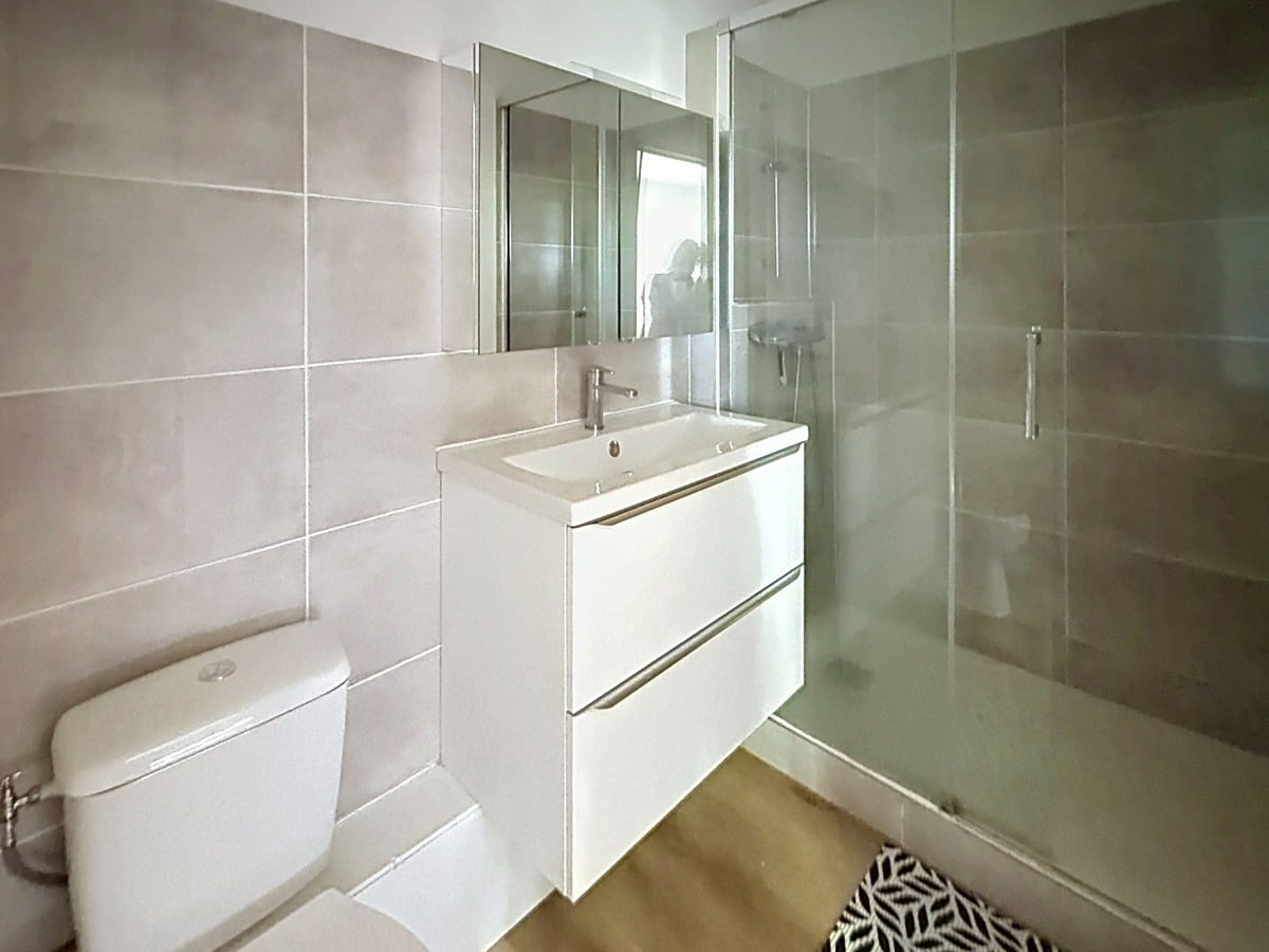 Salle d'eau avec carrelage gris clair, meuble vasque blanc suspendu à deux tiroirs et miroir rectangulaire. Cabine de douche avec paroi vitrée et porte coulissante, WC blanc à gauche, sol en parquet clair.