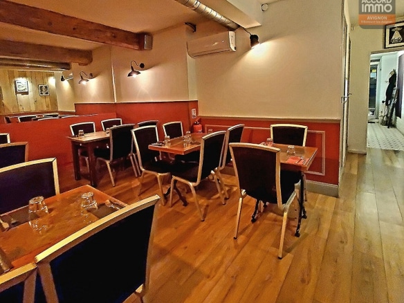 Salle de restaurant avec tables en bois et chaises noires disposées le long d'un mur rouge brique et blanc. Parquet en bois clair, poutres apparentes au plafond, conduit de ventilation métallique et éclairages muraux de style industriel.
