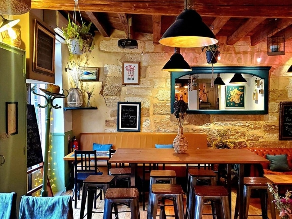 Salle de restaurant avec plafond à poutres apparentes, tables hautes en bois et tabourets métalliques style industriel. Murs en pierre dorée, banquettes en cuir camel, suspensions noires et miroir encadré vert.