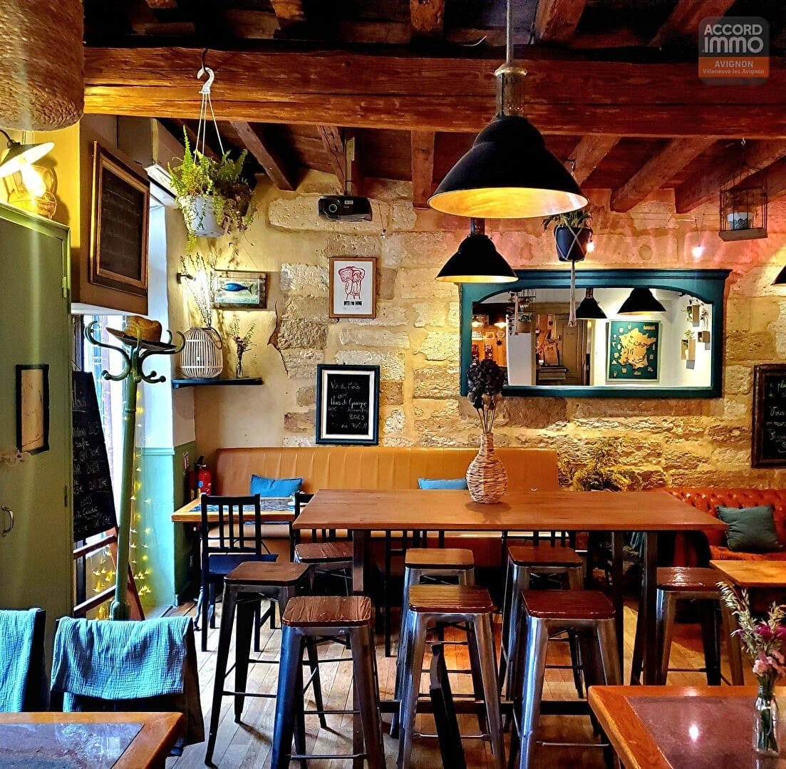 Salle de restaurant avec plafond à poutres apparentes, tables hautes en bois et tabourets métalliques style industriel. Murs en pierre dorée, banquettes en cuir camel, suspensions noires et miroir encadré vert.